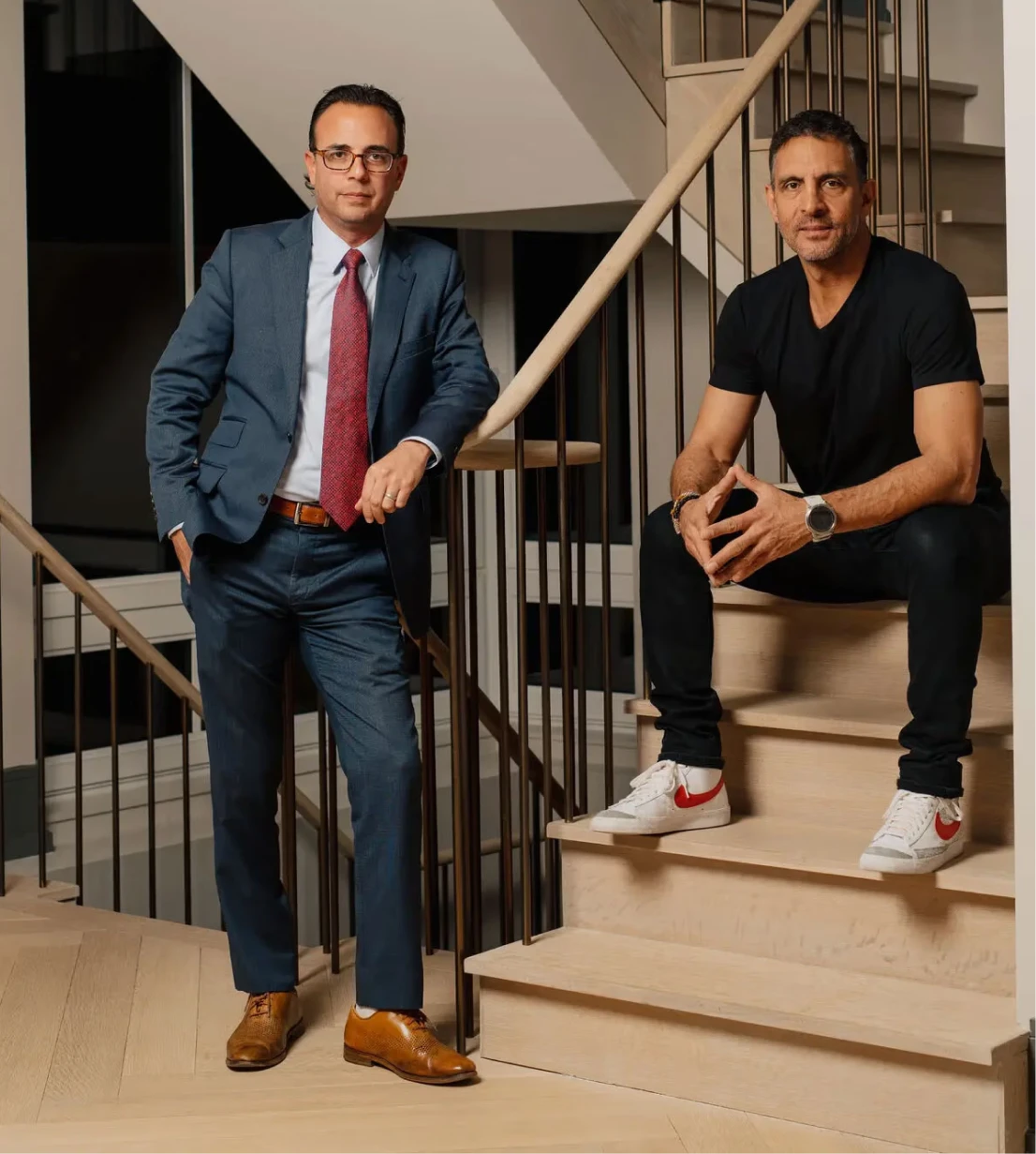 Two men posing on a modern wooden staircase: one standing in a blue suit with a red tie and brown dress shoes, the other seated on the steps in a black t-shirt, black pants and white sneakers with red swooshes.