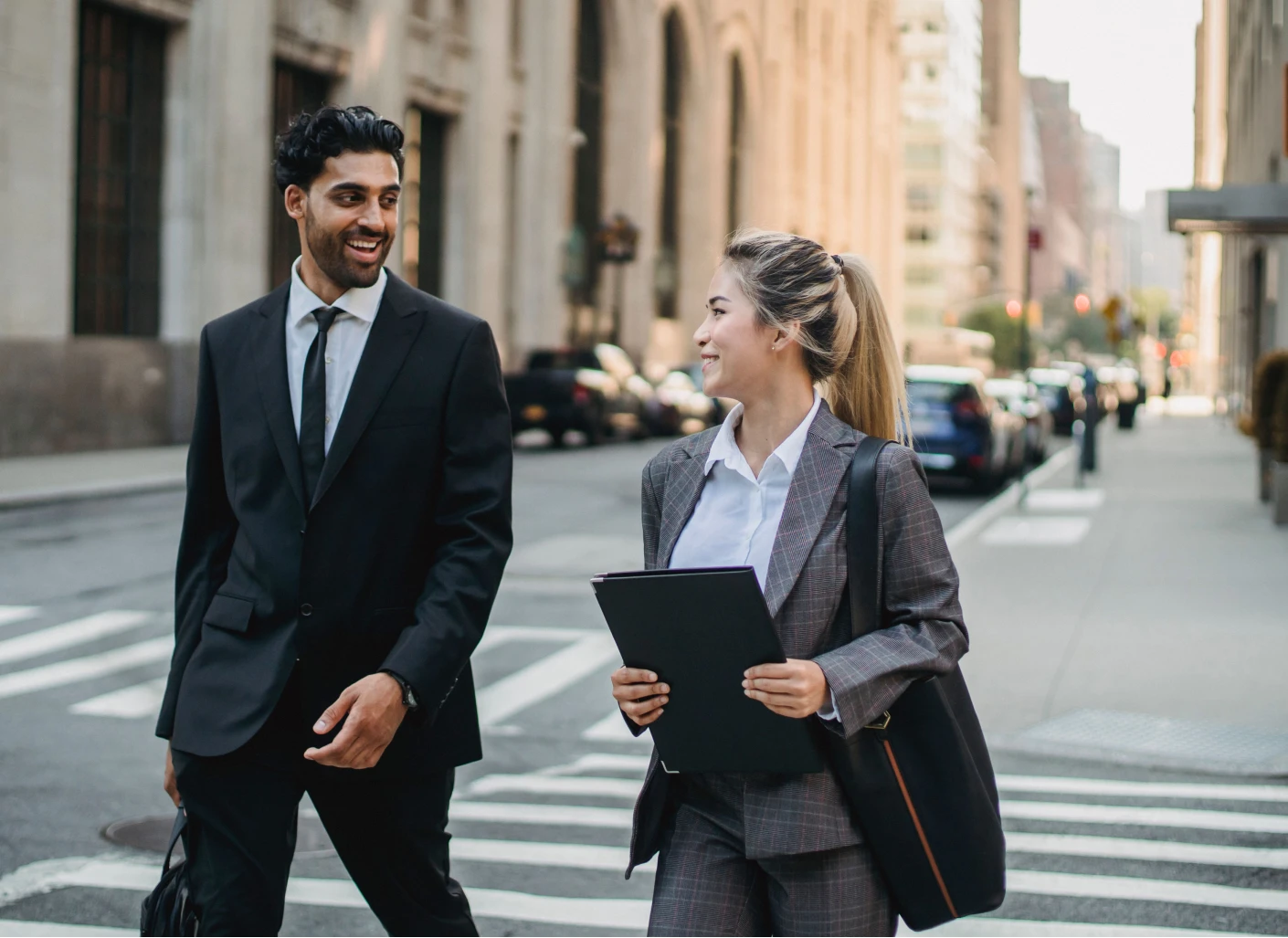 Two business colleagues walking across a city crosswalk, smiling and talking; one holds a portfolio and the other carries a briefcase.